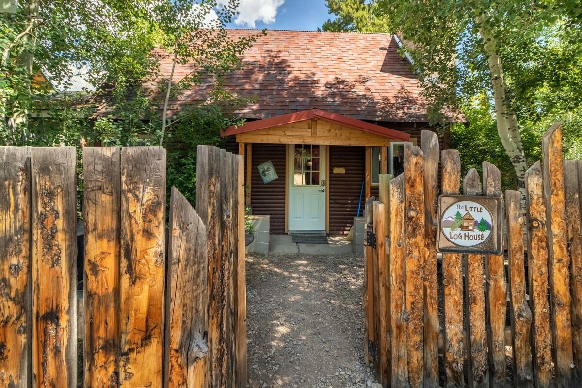 Rustic cabin behind a weathered wooden gate, framed by trees and a gravel path.
