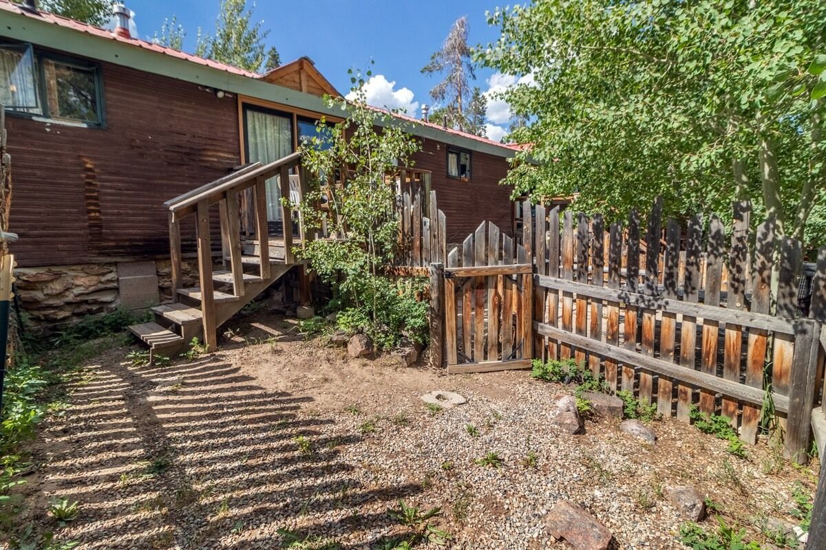 Wooden house with small deck and fenced yard under trees, with dappled sunlight on the ground