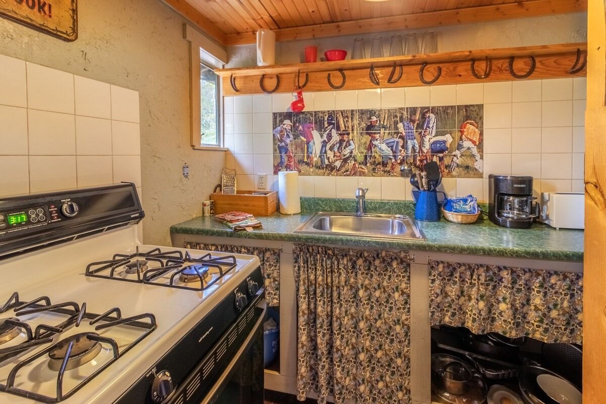 Small kitchen with a gas stove, tiled backsplash, sink, and wooden shelves with hanging mugs.