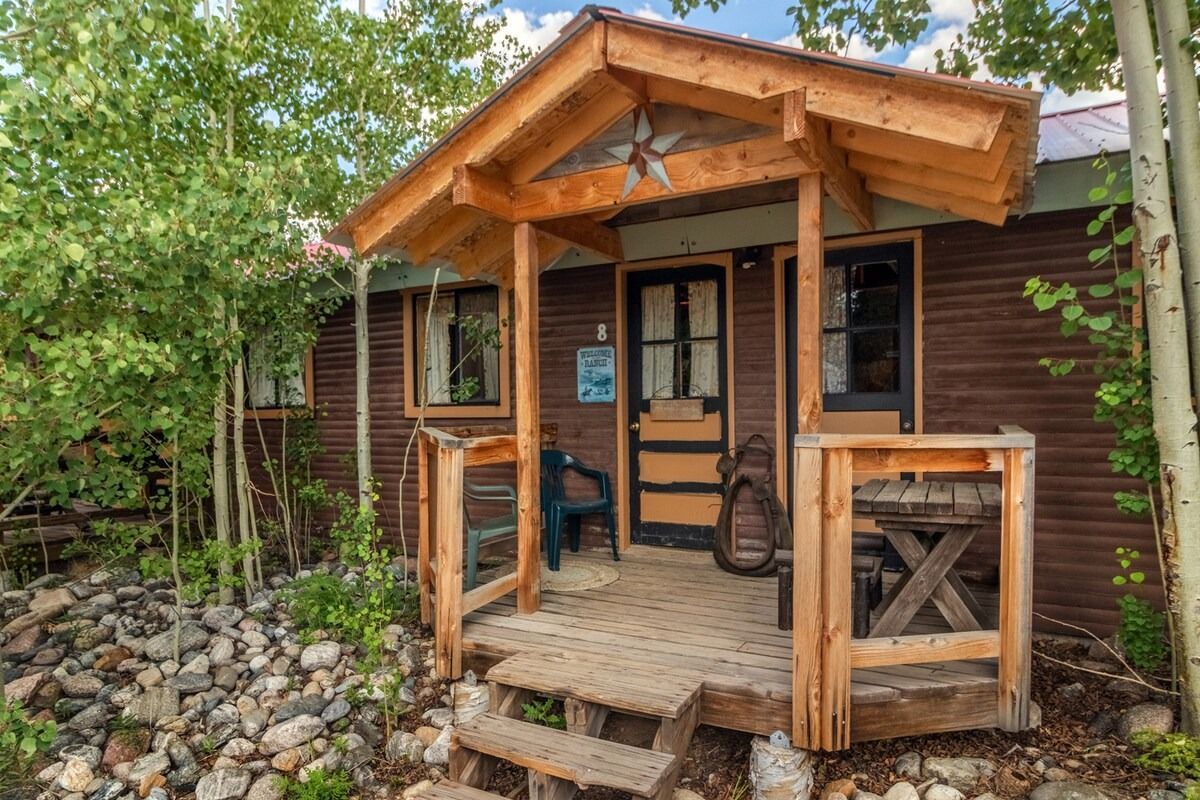 Rustic wooden cabin entrance with a small porch, steps, and rocky landscaping surrounded by greenery