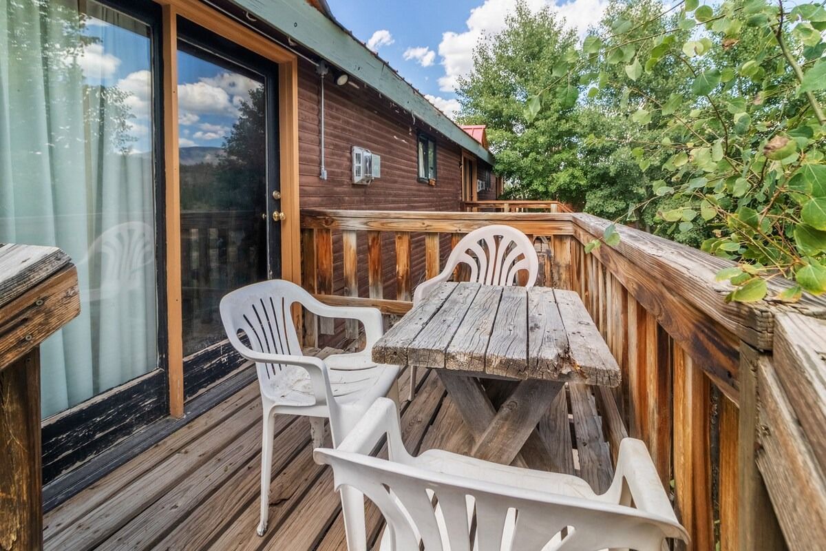Wooden balcony with patio chairs, weathered table, and sliding glass doors overlooking trees