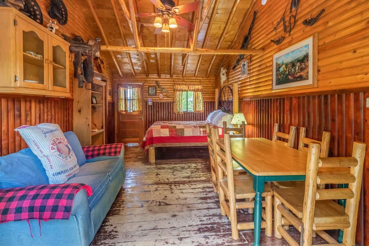 Cozy wooden cabin interior with sofa, dining table, and red bed under a carved wood ceiling