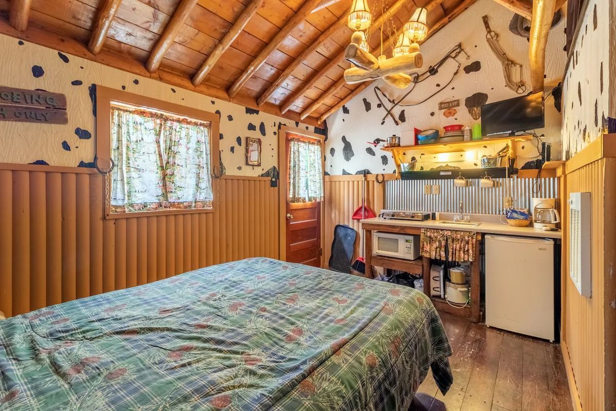 Cozy rustic bedroom with wood-paneled walls, quilted bed, chandelier, and kitchenette area
