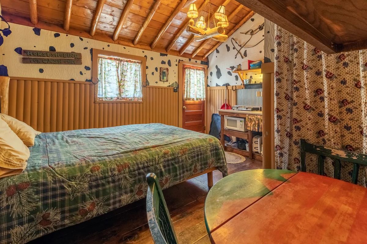 Cozy rustic bedroom with wood-paneled walls, plaid bedspread, and a small round table by the window.