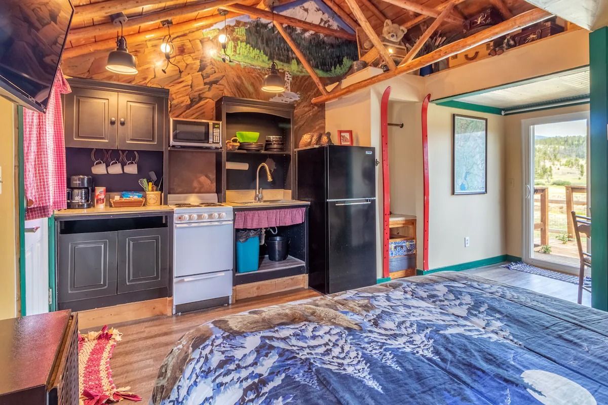 Rustic kitchenette with black cabinets, fridge, and blue rug under exposed wooden beams