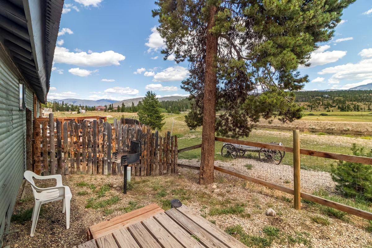 Wooden porch beside fenced yard with white chair, pine tree, and open field under a blue sky