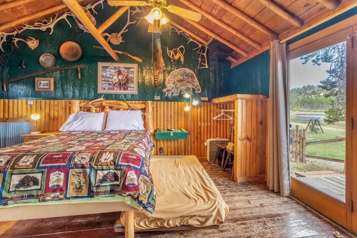 Rustic cabin bedroom with colorful quilt, wood-paneled walls, and a glass door to a grassy outdoor view