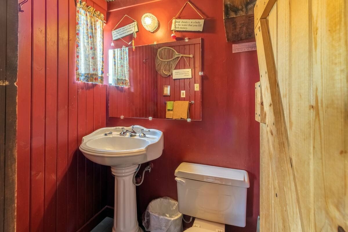 Small red bathroom with pedestal sink, toilet, mirror, and wooden door.