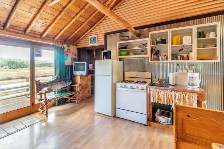 Small rustic kitchen with wood ceiling, stove, fridge, cabinets, and large window overlooking greenery