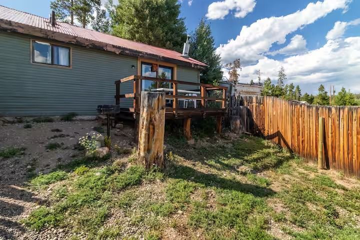 Backyard deck with wooden railings beside a green house, fenced yard, and grassy ground under a blue sky