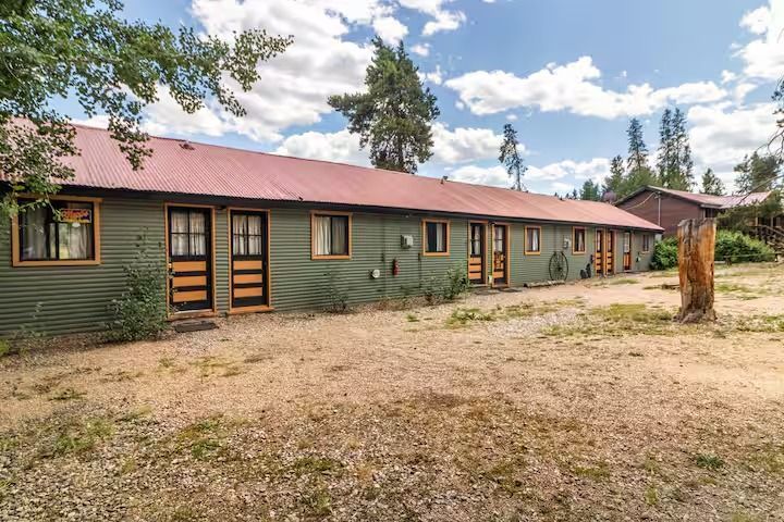 Long green lodge with red roof amid trees and a dirt clearing under a cloudy sky