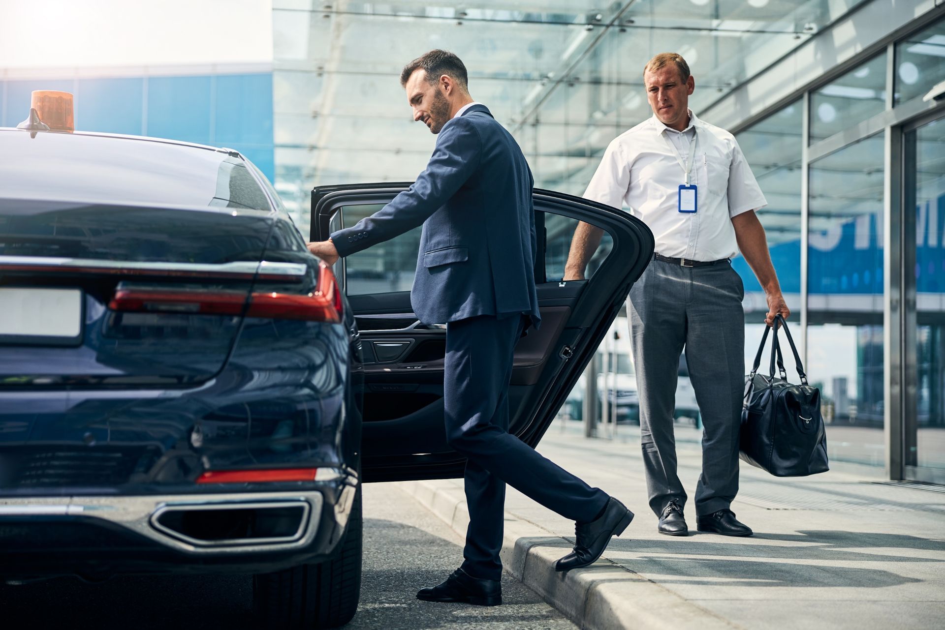 Man in suit exiting a car as another man holds the door open, next to a building.