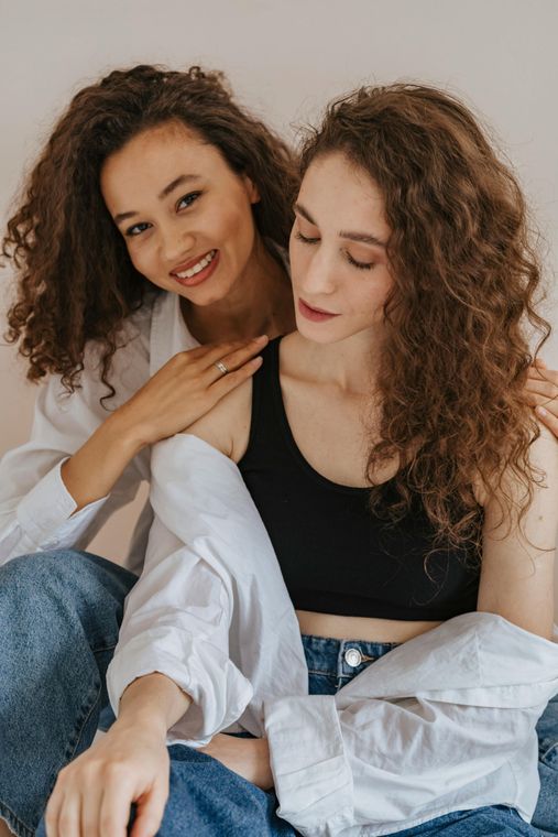 Two women, one smiling with hand on other's shoulder. Both have curly hair and are wearing white shirts and jeans.