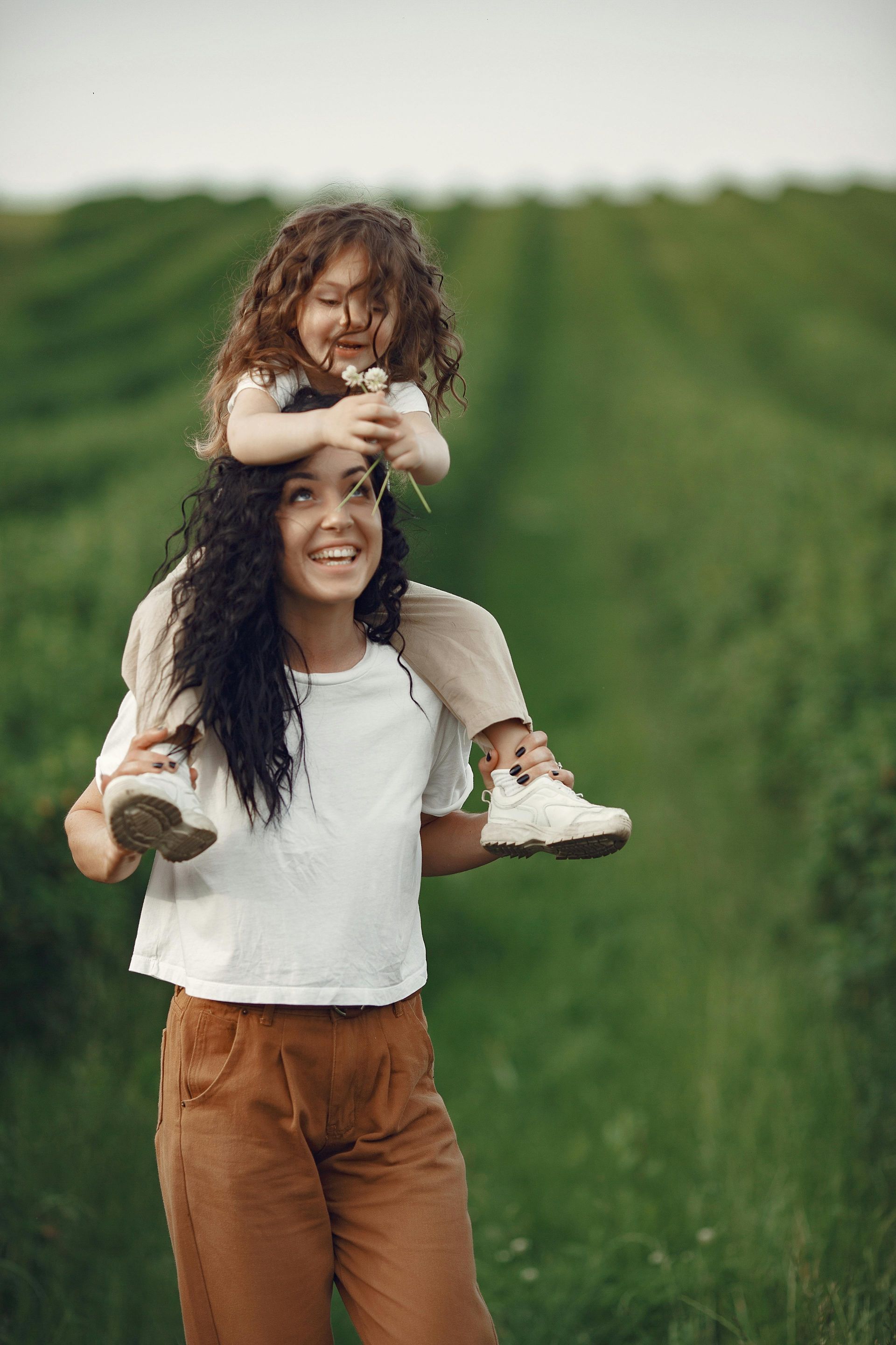 Woman carrying child on shoulders in a green field, both smiling.