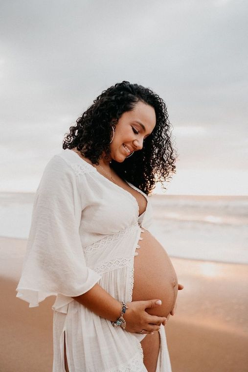 Pregnant person in white top and ripped jeans, touching belly, outdoors in a field with sunlight.