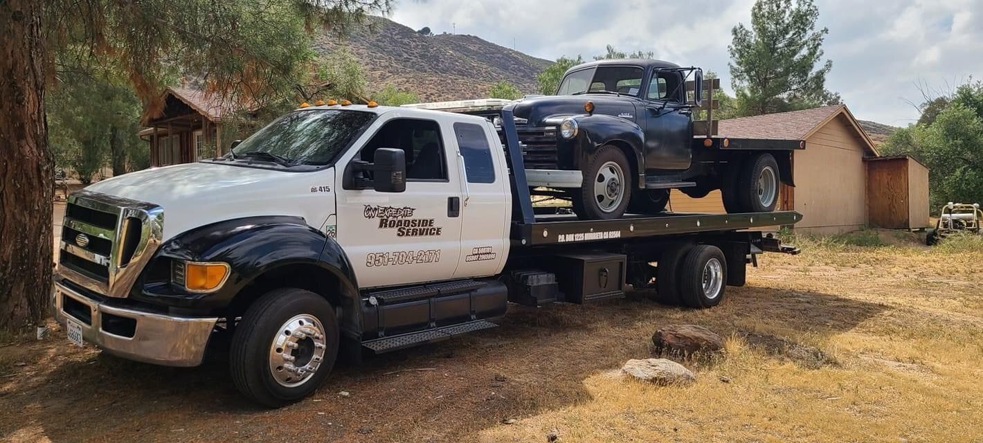 Tow truck carrying a dark pickup in a rural dirt lot beside a small building and trees