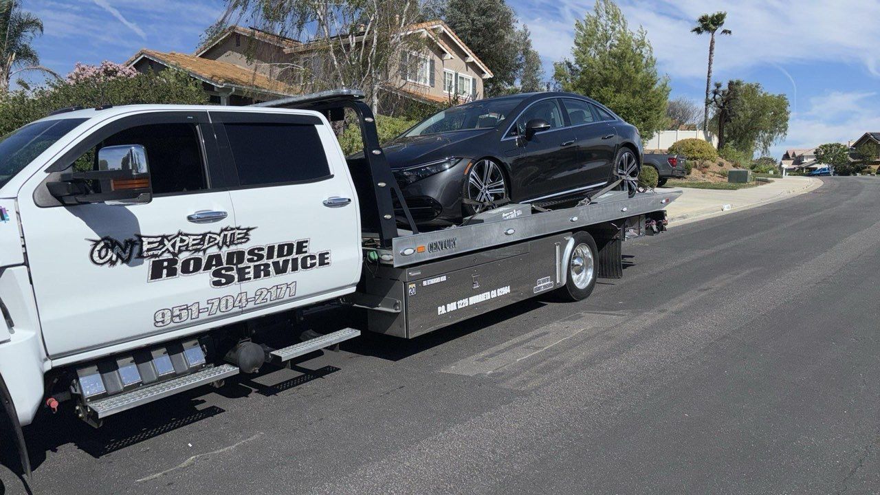 Tow truck hauling a black car on a flatbed beside a suburban street on a sunny day