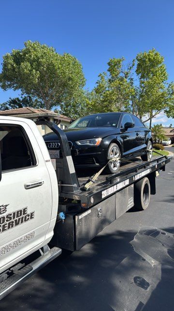 Black SUV on a flatbed tow truck parked on a sunny street with trees in the background