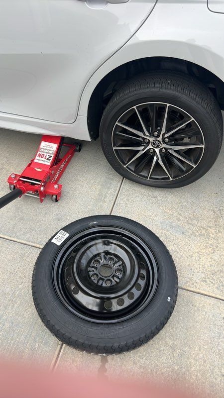 Car with rear wheel removed, jack lifting the vehicle, and spare tire lying on the pavement.