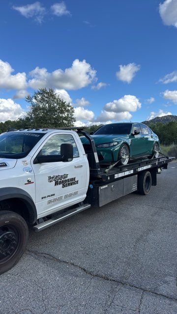 Tow truck hauling a teal car on a flatbed under a blue sky with clouds