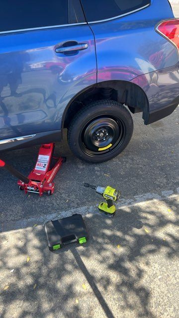Blue SUV lifted by red jack with a wheel removed; two other jacks on the pavement.