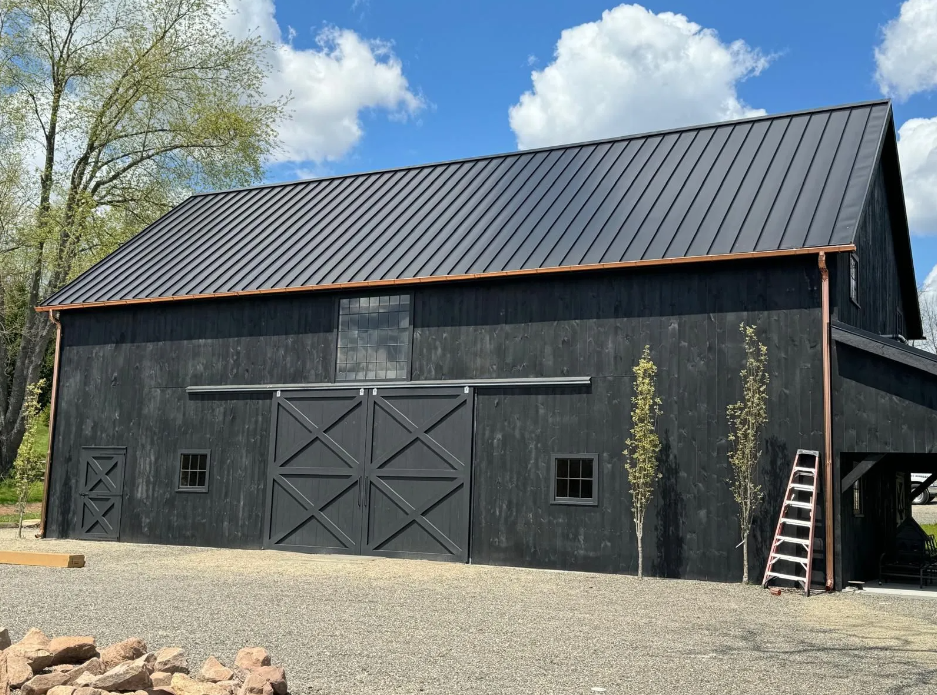 A black barn with a black roof and a ladder in front of it.