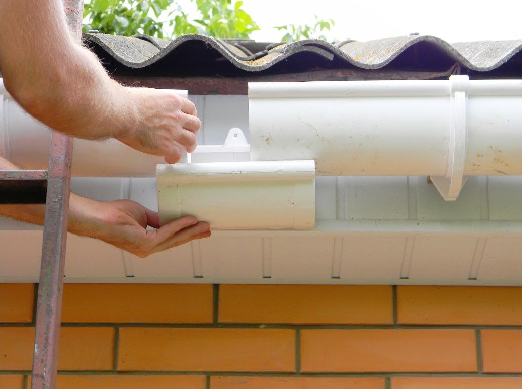 Hands installing white guttering under a tiled roof, with a brick wall below.