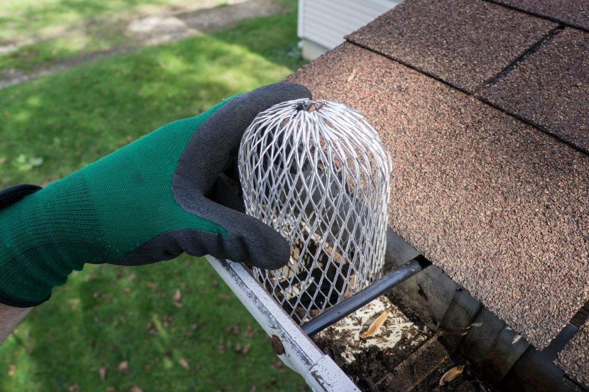 Gloved hand places a mesh gutter guard over a rain gutter beside brown roof shingles.