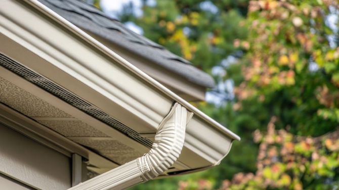White gutter downspout draining along a beige house roof edge with trees in the background