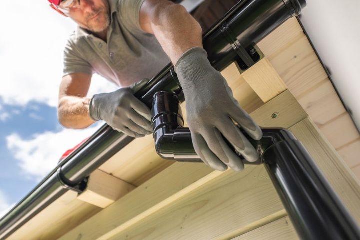 Worker installing black rain gutter on a house roof, wearing gray gloves.