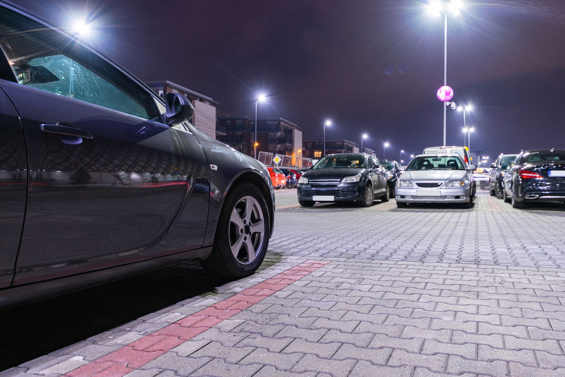 Cars parked on a paved lot at night, lit by tall overhead light poles under a dark purple sky.
