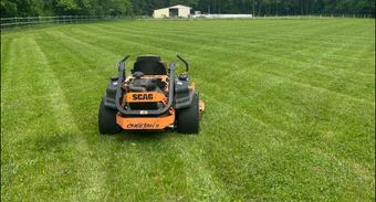 Lawn with fresh mower stripes next to a gravel driveway. Trees and overcast sky.