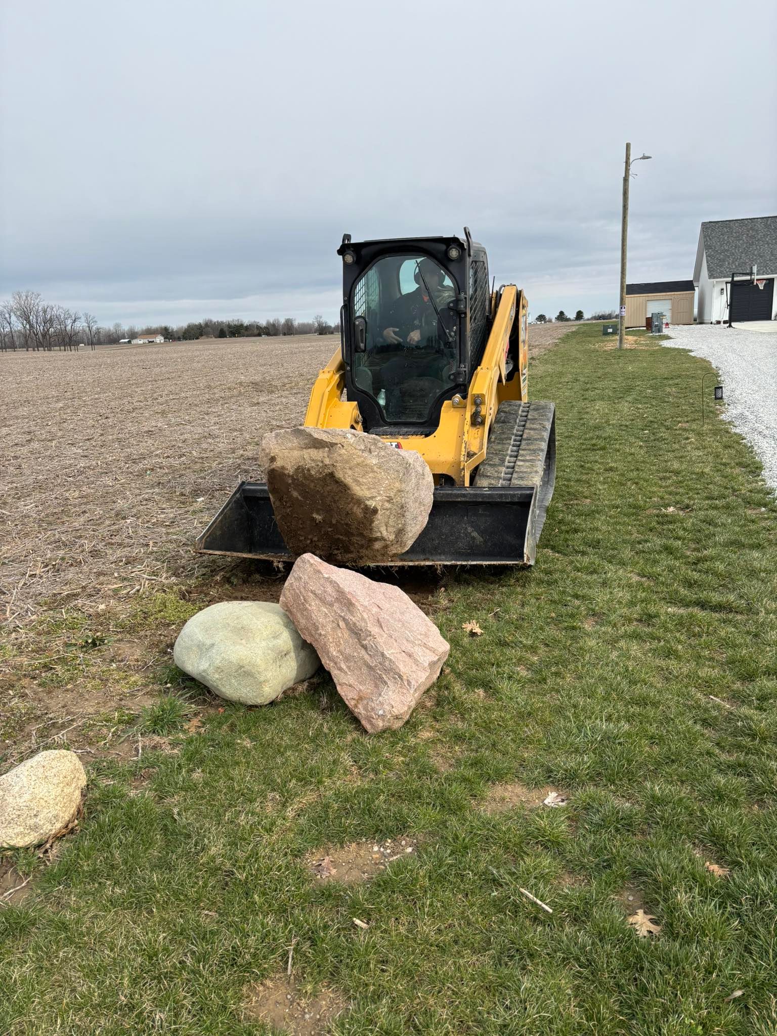 Yellow skid steer loader with a large rock in its bucket, alongside other rocks on a grassy area next to a field.