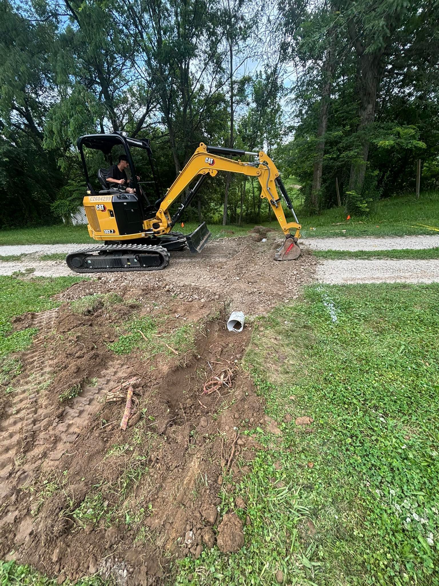 Yellow excavator digging on a gravel driveway, surrounded by grass and trees.