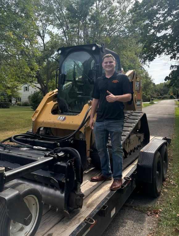 Man giving a thumbs up, standing on a trailer with a yellow track loader.