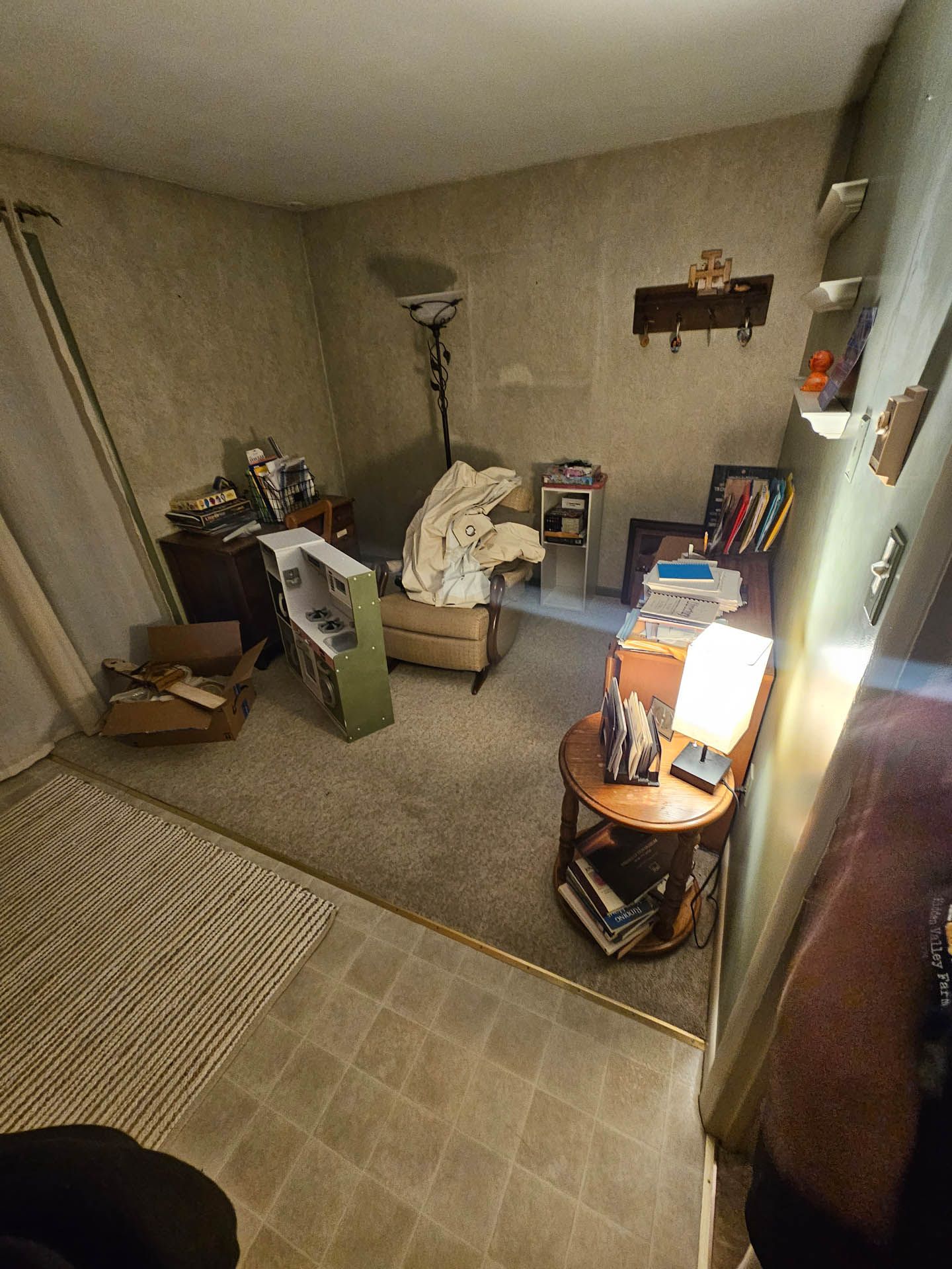 Small cluttered bedroom with bed, lamp, shelves, boxes, and a chair beside a window.