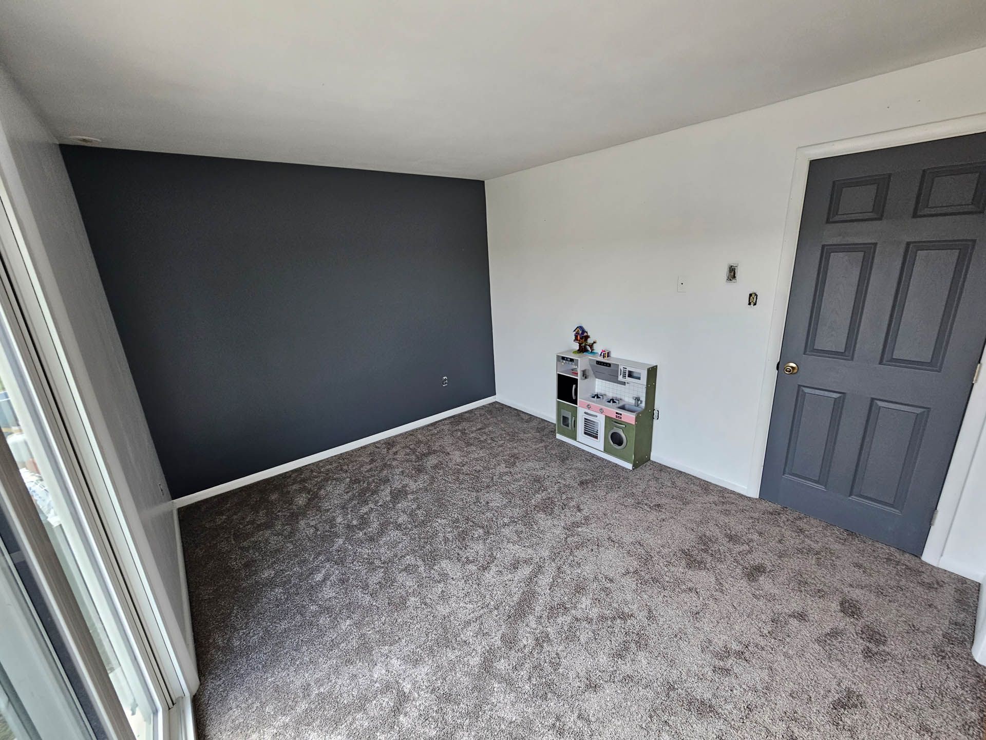 Empty bedroom with gray carpet, dark accent wall, white walls, and a closed gray door.