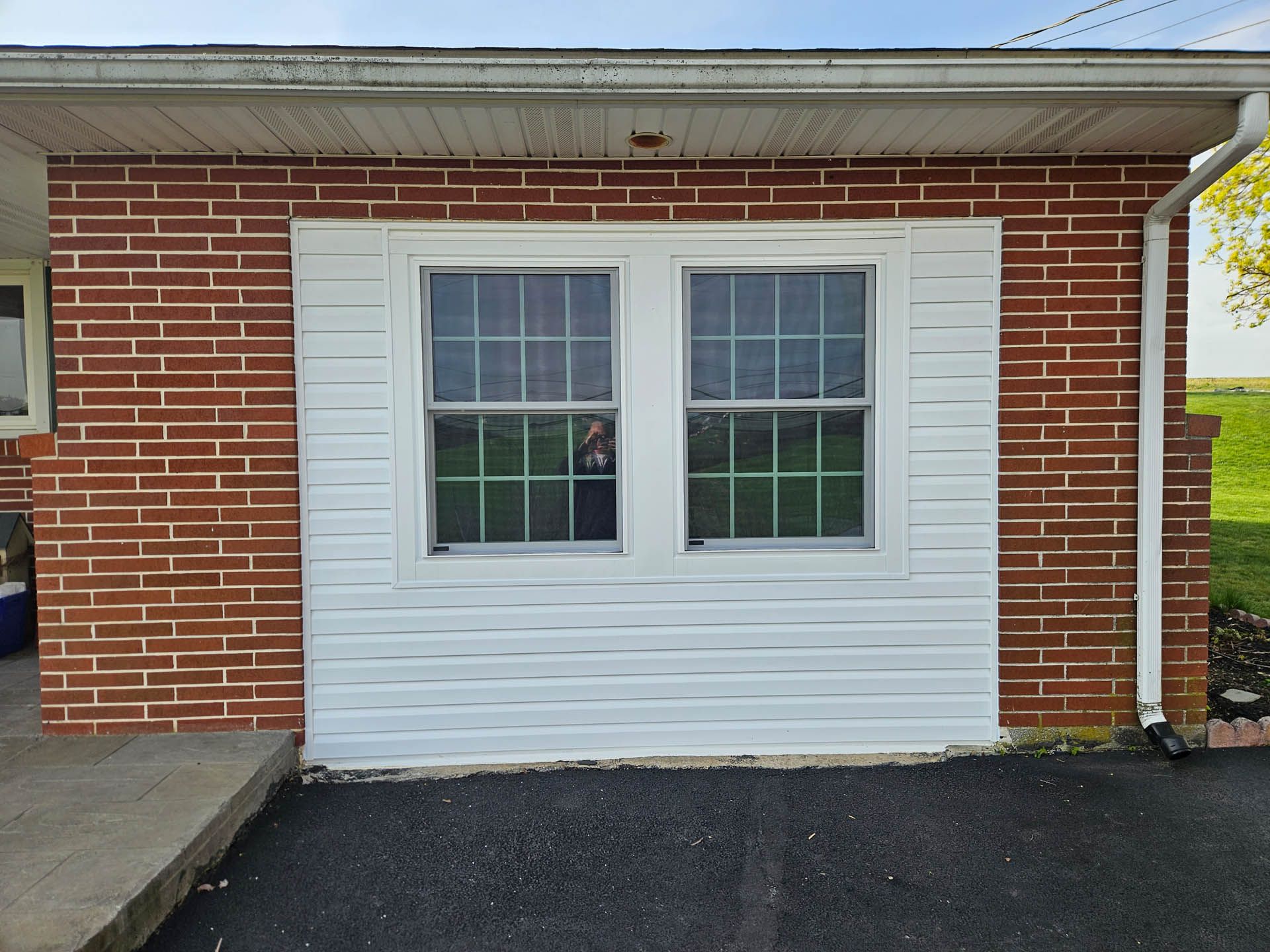 Brick building wall with white-framed double window and shutters beside a concrete walkway