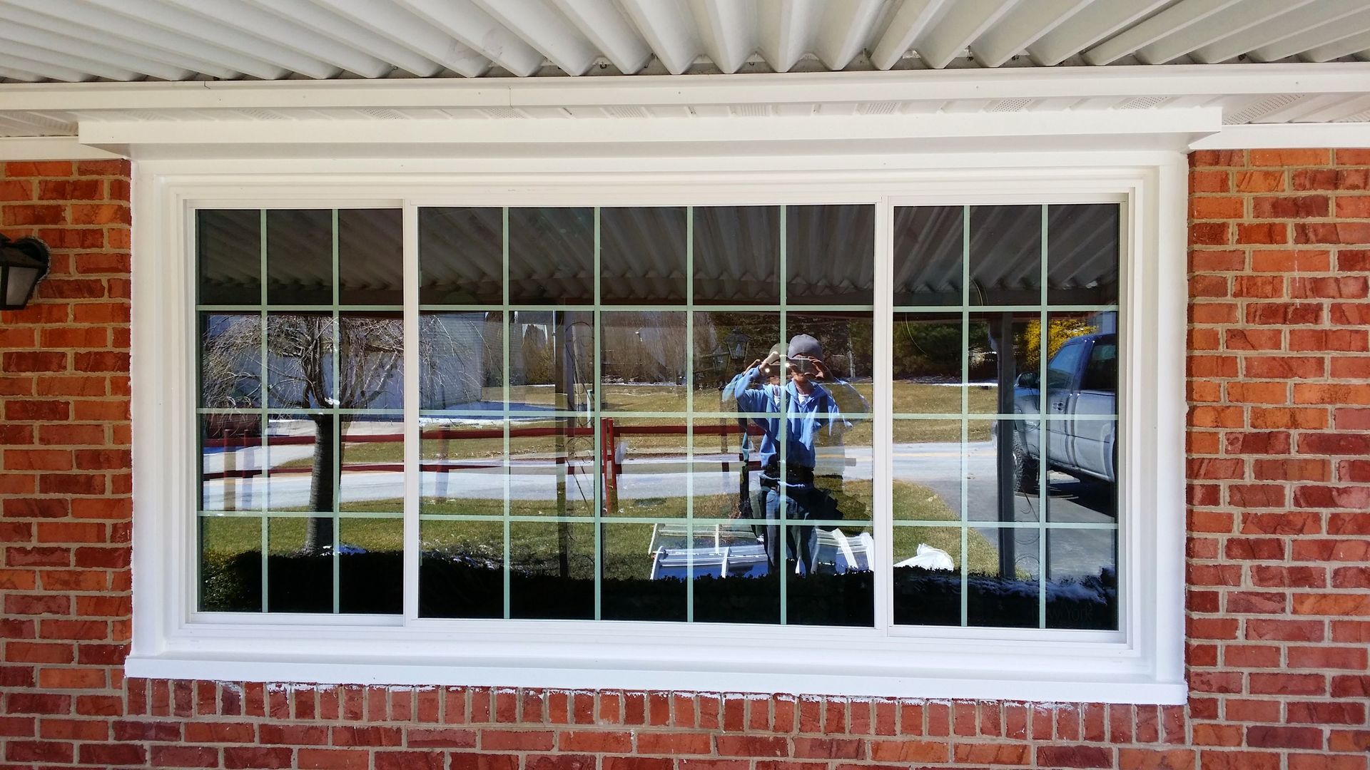 A white-framed sliding window with a grid pattern, installed in a red brick wall, reflecting a person taking a photo.