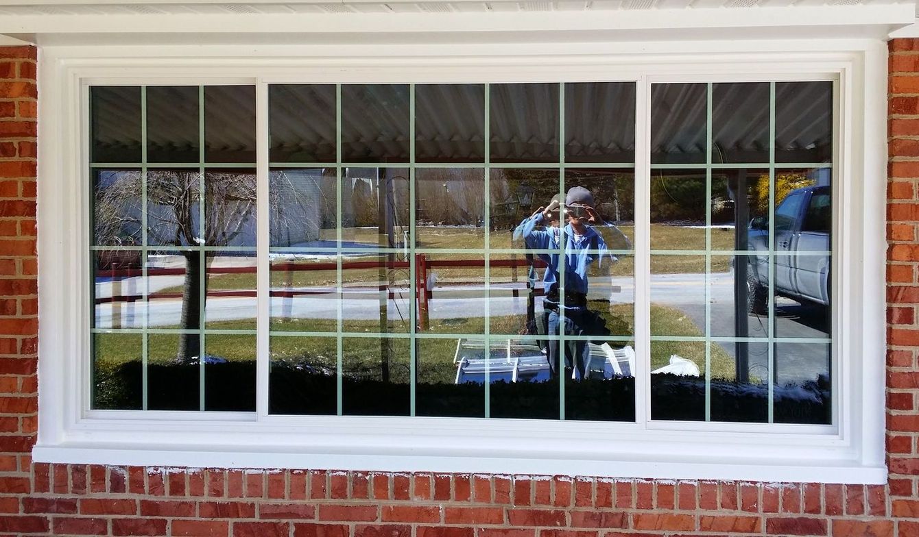 A white-framed window with a grid pattern, reflecting a person outdoors and a parked vehicle in a brick-walled setting.