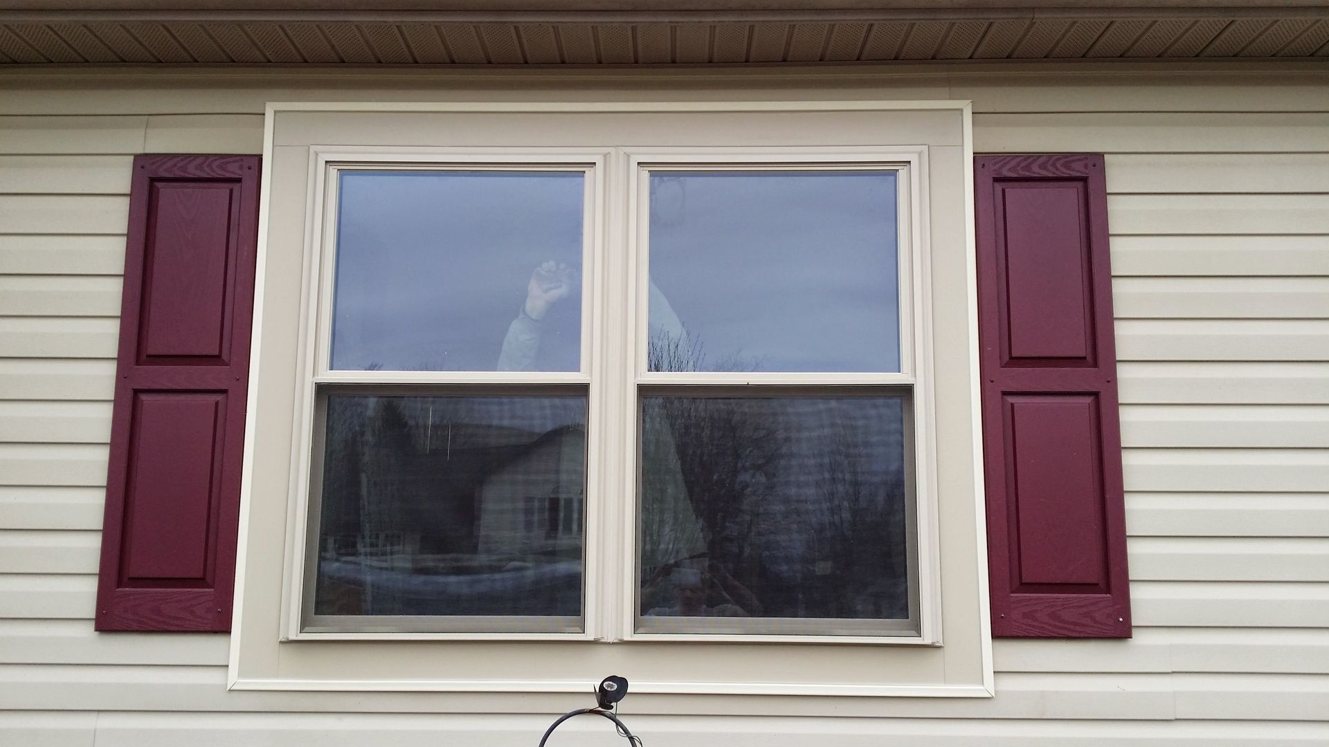 A hand waves from inside a house through a double-pane window flanked by maroon shutters on light-colored vinyl siding.