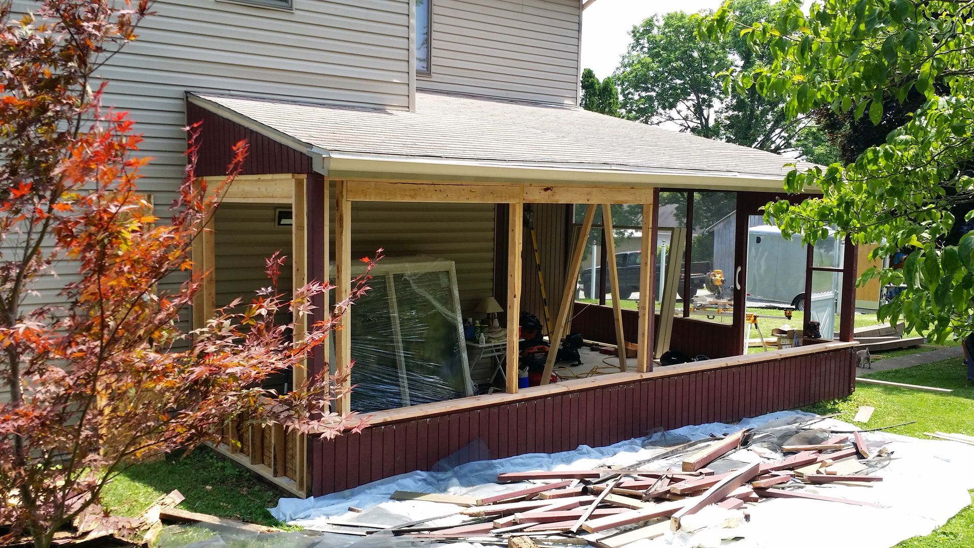 An unfinished wooden porch addition attached to the side of a house with maroon siding and light-colored shingles.