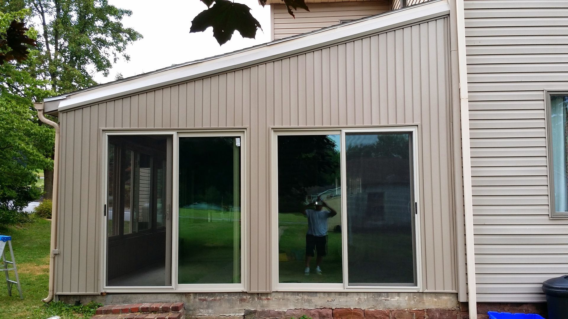 Exterior view of a beige home with two sliding glass doors and a slanted roof, surrounded by green trees and grass.