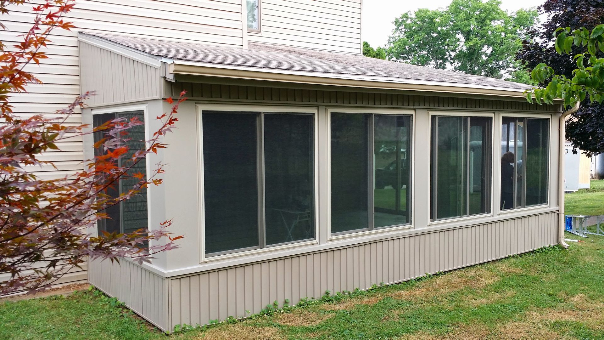 A beige sunroom addition with sliding windows and a slanted, corrugated roof attached to the side of a house.
