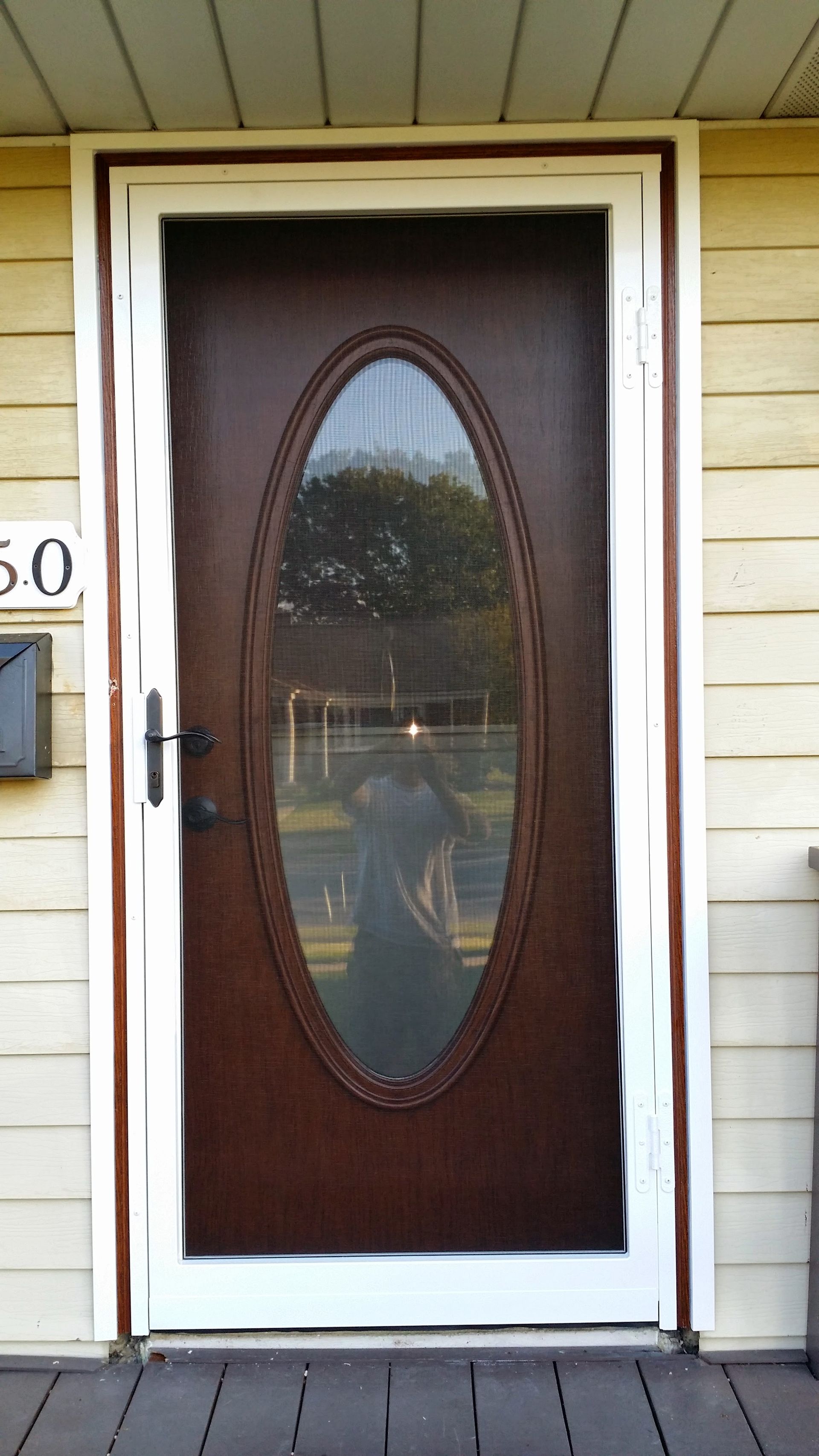 A brown storm door with a central oval glass pane, set in a white frame on a yellow-sided house.