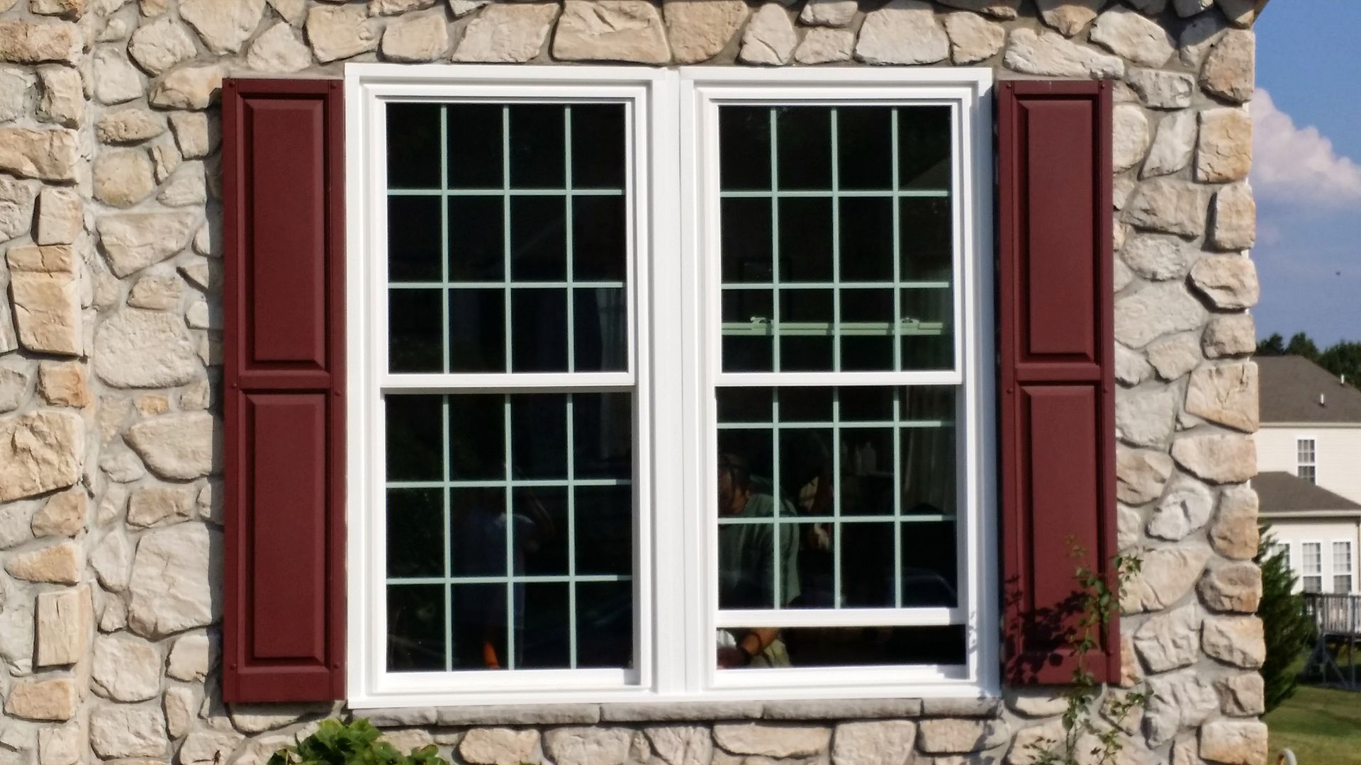 A double-hung white window with a grid pattern, flanked by dark red shutters, set into a stone wall.