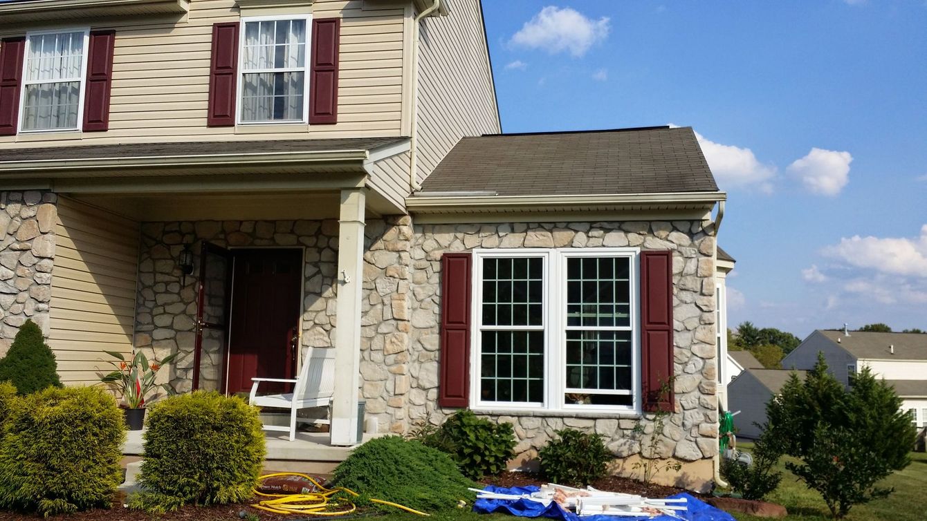 Tan-sided suburban house with a stone front, dark red shutters, a porch, and a blue tarp on the front lawn.