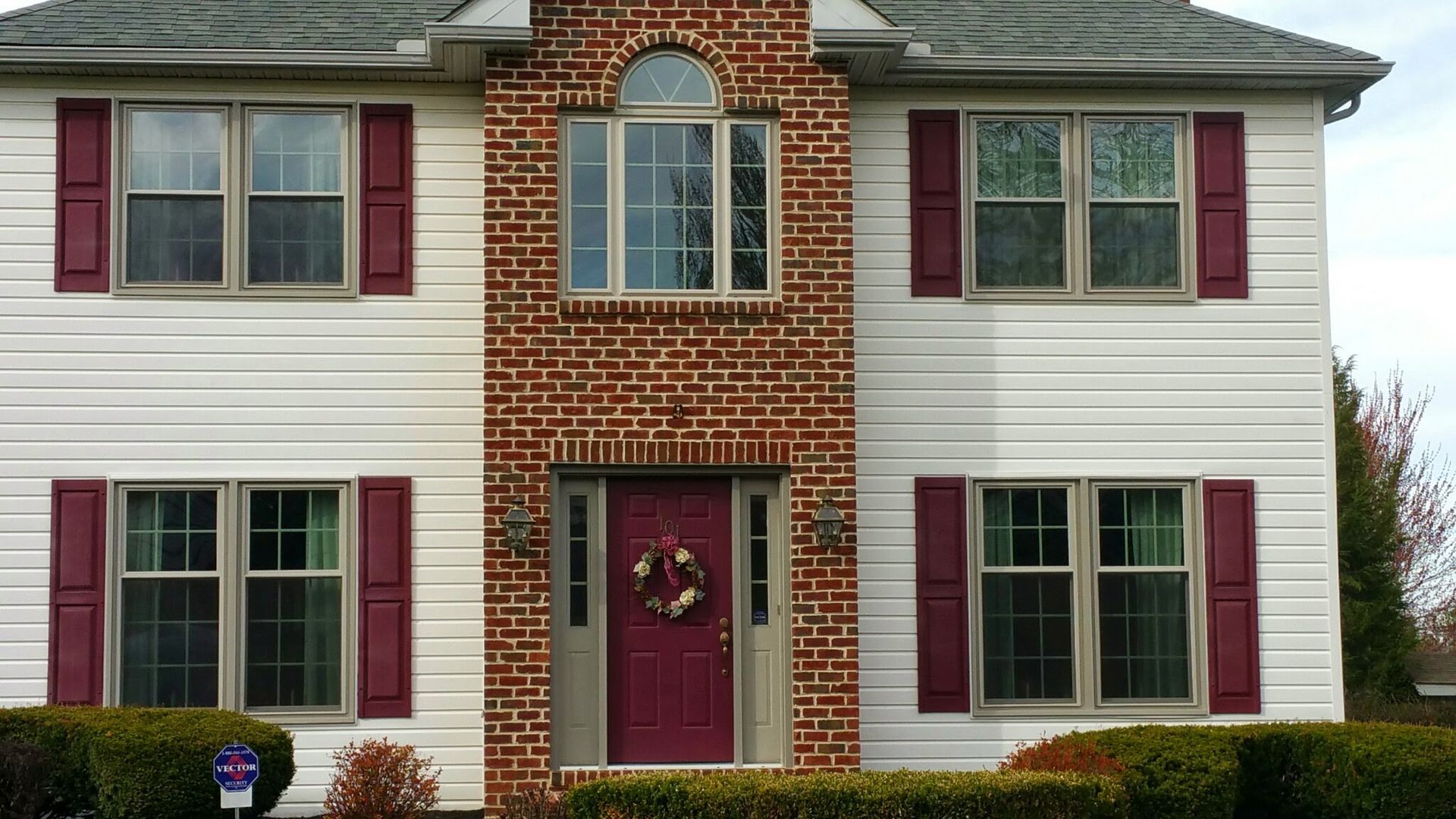 A two-story house with white siding, red shutters, and a central brick facade featuring a dark red front door.