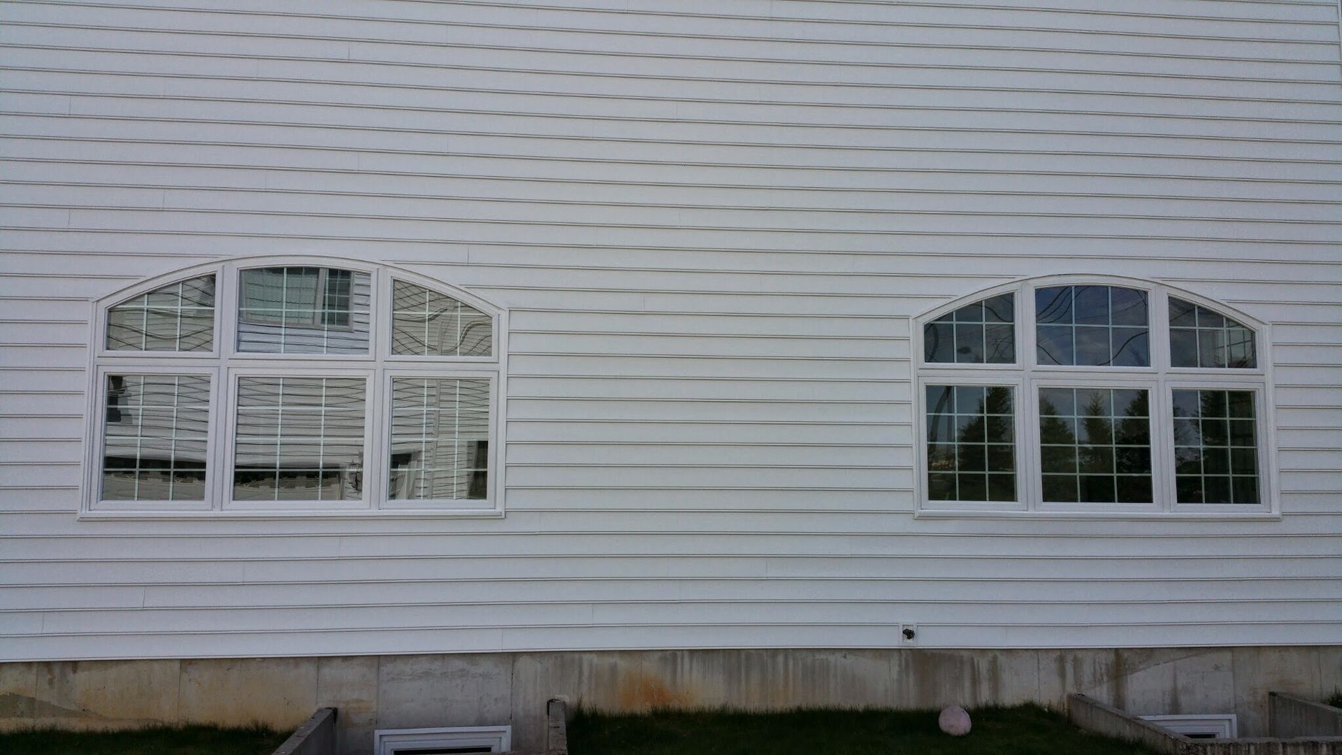 White siding on a building wall with two arched, multi-pane windows and a concrete foundation below.