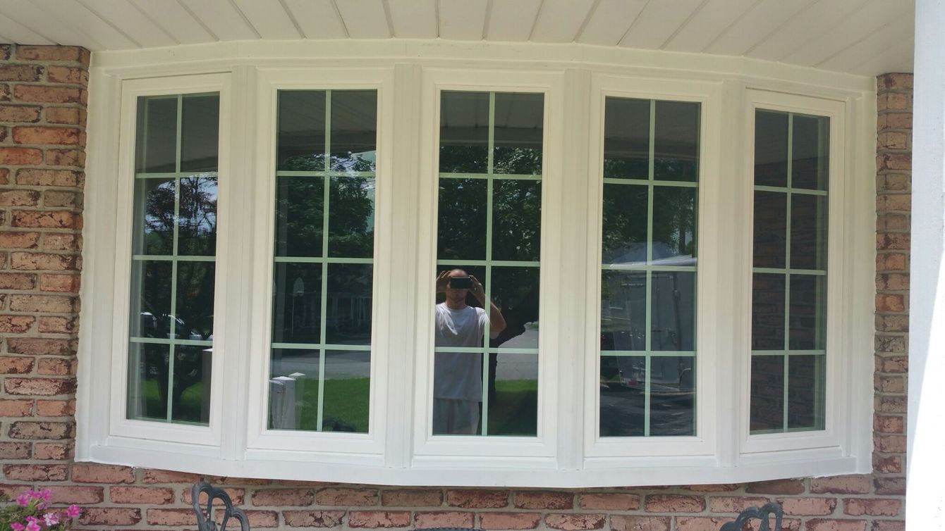 A five-panel white bay window installed in a brick exterior wall, reflecting a person standing outside.